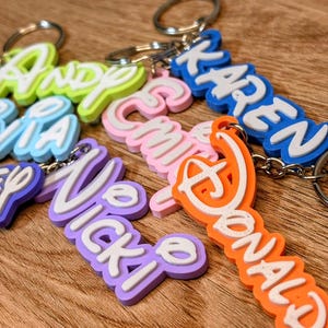 May include: A collection of custom 3D-printed keychains featuring names in various colours including neon green, light blue, purple, pink, and orange, each with white lettering and attached to a silver-toned metal split ring for keys.