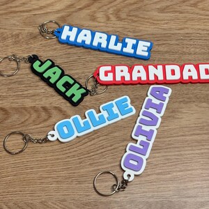 May include: Five rectangular silicone keychains with metal rings, each featuring raised white lettering. The names displayed are HARLIE in blue, JACK in green, GRANDAD in red, OLLIE in light blue, and OLIVIA in purple, all in a bold block font.