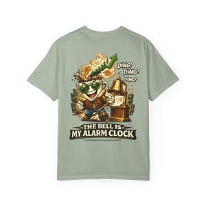 May include: A sage green t-shirt featuring a graphic of a coffee mug character wearing sunglasses and a jacket, ringing a brass bell. Above the mug is a green upward-trending stock chart. Text reads: DING! DING! DING! THE BELL IS MY ALARM CLOCK.