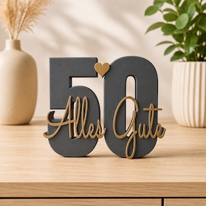 Puede incluir: Una decoración de pie con el número 50 en gris oscuro, que presenta un pequeño corazón dorado entre los dígitos y el texto Alles Gute en cursiva dorada superpuesto en la parte frontal. La pieza tiene un acabado mate y se mantiene en pie.