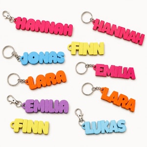 Könnte beinhalten: Eine Sammlung bunter, 3D-gedruckter Schlüsselanhänger mit Namen in Blockbuchstaben. Die Namen umfassen Hannah in Pink, Jonas und Lukas in Hellblau, Finn in Gelb, Lara in Orange sowie Emilia in Lila und Pink, jeweils mit Metallring oder Karabiner.