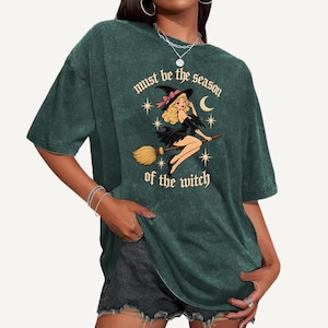May include: A dark green mineral-washed t-shirt featuring a graphic of a blonde witch in a black dress and hat riding a broomstick. The text "must be the season of the witch" is printed in a cream-colored font, surrounded by stars and a crescent moon.