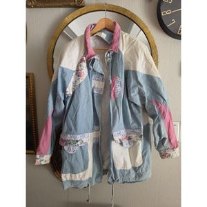 May include: A long-sleeve jacket featuring a color-blocked design in light blue, white, and pink. The garment includes panels with a floral pattern, a high collar, a front zipper closure, and two large patch pockets on the lower front section.
