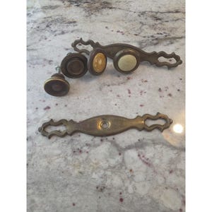 Può includere: Una collezione di ferramenta color ottone che include due maniglie per cassetti allungate con intagli decorativi, due pomelli rotondi con bordi marrone scuro e un pomello rotondo con inserto centrale color crema e bordo marrone scuro.