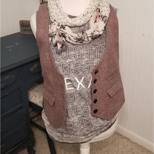 May include: A sleeveless brown herringbone vest with five black buttons and two front pockets worn over a gray tank top with the white text AUSTIN TEXAS repeated vertically. A white scarf with a black polka dot and floral pattern is draped around.