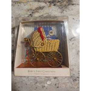 May include: A beige wicker-style baby carriage ornament with gold-tone metal wheels and a red accent, encased in a clear plastic box. The packaging features the text Baby's First Christmas Dated Ornament printed on the front base of the display.