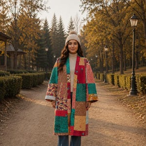 May include: A long, open-front coat featuring a colourful patchwork design with various floral and geometric patterns in shades of teal, red, brown, and yellow. The coat is worn over a beige turtleneck sweater, blue jeans, and a beige knit beanie.