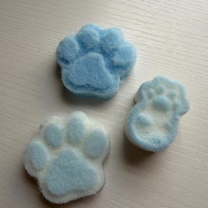 May include: Three paw-shaped objects made of a fuzzy, light blue and white material. Two of the objects are larger and rounder, featuring a distinct central pad and four smaller toe pads, while the third is smaller and more elongated in shape.