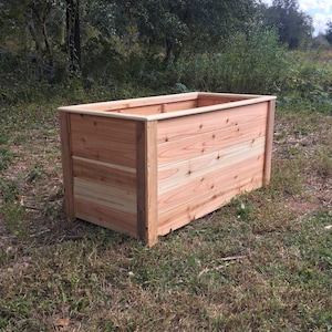 May include: A rectangular planter box constructed from horizontal cedar wood planks with a smooth top rim. The structure features vertical corner posts and a natural light brown wood grain finish, showcasing a sturdy, box-like design for gardening.