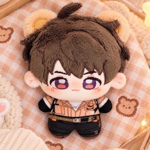 May include: A plush doll featuring a character with short brown hair and bear ears. The doll has purple eyes, a tan collared shirt with black accents, and black pants. The plush has a soft texture and a small tuft of hair on top of its head.