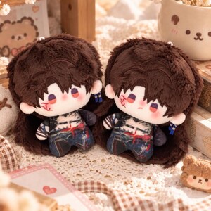May include: Two plush dolls featuring dark brown fuzzy hair, pale skin with red facial markings, and dark eyes. Each doll wears a dark patterned outfit with blue and red accents, and small blue and white dangling accessories on the sides of their heads.