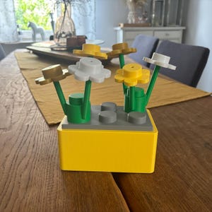 Könnte beinhalten: Ein rechteckiger gelber Sockel mit einer grauen Oberseite, die vier kreisförmige Noppen aufweist. Vier Kunststoffblumen mit grünen Stielen sind am Sockel befestigt. Die Blüten haben weiße, gelbe und goldfarbene Blütenblätter mit kleinen runden Mitten.