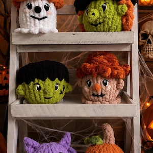 May include: Six plush crochet figures arranged on a white wooden shelf. The collection includes a white ghost with an orange hat, a green witch with a black and purple hat, a green monster, a tan scarecrow, a purple bat, and an orange pumpkin.