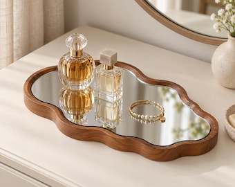 May include: An irregular, cloud-shaped vanity tray featuring a smooth, dark wood frame and a reflective mirrored surface. The tray holds two glass perfume bottles with gold accents and a delicate gold-toned beaded bracelet.
