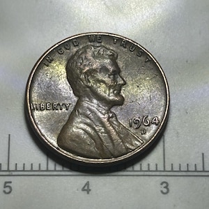 以下が含まれることがあります： 虹色のトーンを持つ1964年Dミントマークのリンカーン1セント硬貨。エイブラハム・リンカーンの横顔、IN GOD WE TRUST、LIBERTY、1964の刻印があり、直径1.9cmを示す定規の上に配置されています。