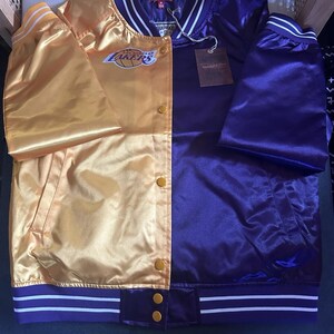 May include: A satin varsity jacket featuring a split design with a gold left side and a purple right side. The jacket includes a Los Angeles Lakers logo on the chest, a striped ribbed collar and cuffs, and gold snap buttons down the front center.