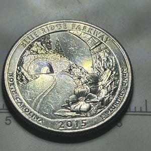 May include: A silver-coloured 2015 North Carolina Blue Ridge Parkway quarter coin featuring a stone tunnel, a winding road, and dogwood flowers. The text reads BLUE RIDGE PARKWAY, NORTH CAROLINA, 2015, and E PLURIBUS UNUM. A ruler is visible at the bottom.