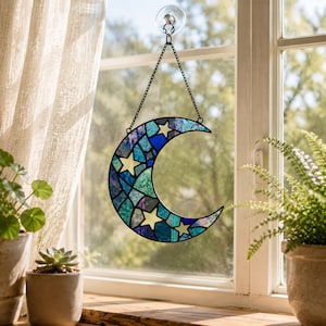 May include: A crescent moon suncatcher crafted from stained glass in shades of blue, teal, and purple, featuring embedded pale yellow star shapes. The piece is suspended by a black metal chain attached to a clear suction cup on a window pane.