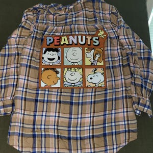 May include: A long-sleeve button-down shirt featuring a tan, blue, and white plaid pattern. A large rectangular patch on the back displays the text PEANUTS above a grid of six illustrations depicting characters including Snoopy and Charlie Brown.