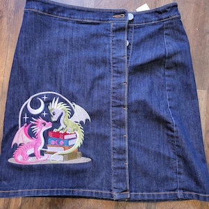 May include: Dark wash denim skirt with a button-up front. The skirt features an embroidered design of two dragons, one pink and one green, perched on books under a crescent moon and stars.