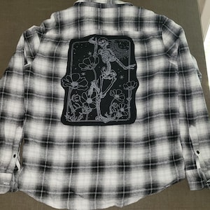 May include: A long-sleeved button-down shirt featuring a black and white plaid pattern. The back of the shirt displays a large rectangular black patch with a white line-art illustration of a skeleton dancing among flowers, stars, and a sun.