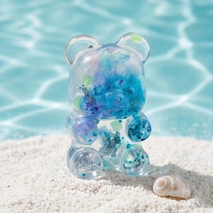 May include: A clear resin bear figurine filled with blue, purple, and green glitter and confetti-like inclusions. The bear has a smooth, rounded shape and is positioned next to a small, spiralled white seashell on a textured white surface.