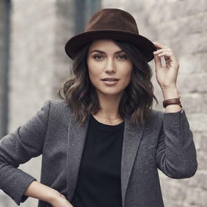 Könnte beinhalten: Ein dunkelbrauner Filz-Fedora-Hut mit strukturierter Krempe, getragen zu einem strukturierten anthrazitfarbenen Blazer über einem schlichten schwarzen T-Shirt. Eine Armbanduhr mit braunem Lederarmband ergänzt die neutralen Töne des Outfits.