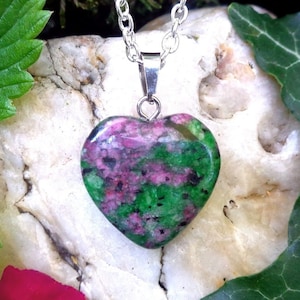 May include: A heart-shaped pendant necklace featuring a green and pink gemstone. The pendant is on a silver chain and is resting on a rock with green leaves.