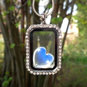 May include: A silver locket necklace with a clear glass heart-shaped charm inside. The charm is filled with a light blue substance. The locket is surrounded by small clear rhinestones.