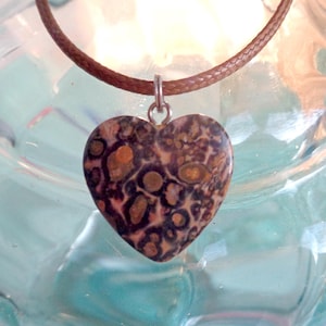 Puede incluir: Un collar con colgante en forma de corazón. El colgante es de piedra moteada con colores marrón, beige y crema. Cuelga de un collar de cordón marrón. El colgante está suspendido de una fianza de color plateado.