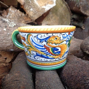 Könnte beinhalten: Eine Keramiktasse mit einem farbenfrohen Drachendesign. Die Tasse hat einen grünen Henkel und Boden, mit einem gelben und orangen Rand. Der Korpus ist weiß mit blauen, gelben und orangen Dekorelementen. Die Tasse steht auf einer Holzoberfläche.