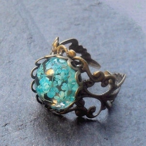 May include: A ring with a delicate, ornate design featuring a round, clear glass cabochon filled with tiny blue flowers. The ring is made of bronze and has an adjustable band.