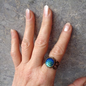 Puede incluir: Un anillo con un dise&ntilde;o de escamas de sirena azul y verde iridiscente en una banda de filigrana de bronce.