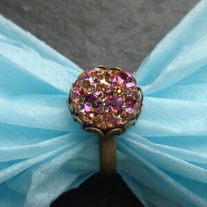 Puede incluir: Un anillo con una gran piedra preciosa redonda, iridiscente rosa y dorada, engastada en un engaste de color bronce. El anillo est&aacute; sobre un fondo azul claro.