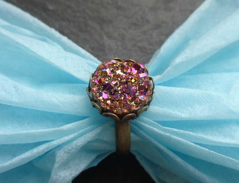 Puede incluir: Un anillo con una gran piedra preciosa redonda, iridiscente rosa y dorada, engastada en un engaste de color bronce. El anillo est&aacute; sobre un fondo azul claro.