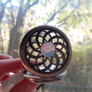 May include: Antique bronze-colored pocket watch with an openwork design. The watch face features a central, iridescent, round stone. The watch is held open, revealing the inner workings. The background is a blurred view of trees.