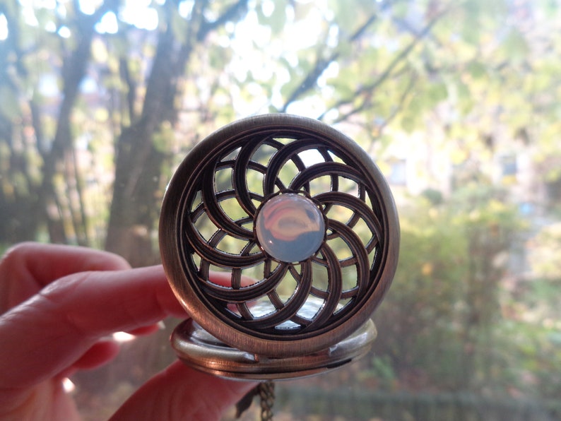 May include: Antique bronze-colored pocket watch with an openwork design. The watch face features a central, iridescent, round stone. The watch is held open, revealing the inner workings. The background is a blurred view of trees.