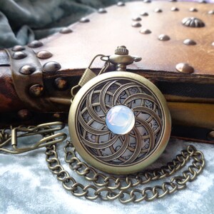 May include: Antique brass pocket watch with a decorative face featuring a swirling pattern and a central opalescent stone. The watch is attached to a chain and rests on a soft, textured surface, with a leather-bound book in the background.