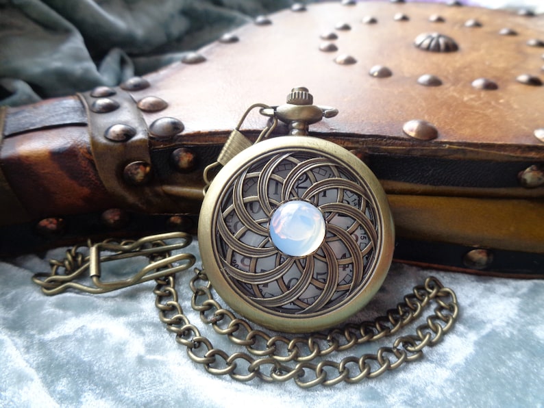 May include: Antique brass pocket watch with a decorative face featuring a swirling pattern and a central opalescent stone. The watch is attached to a chain and rests on a soft, textured surface, with a leather-bound book in the background.