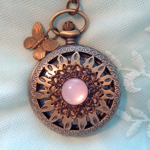 May include: A vintage-style pocket watch with a delicate butterfly charm and a pink face. The watch has a detailed, ornate design with a circular, openwork pattern.