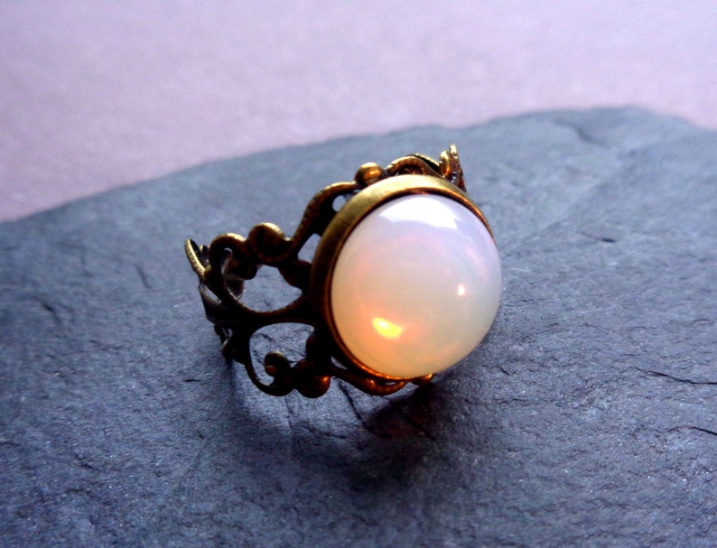 Puede incluir: Un anillo de &oacute;palo blanco con un engaste de lat&oacute;n adornado. El anillo tiene un dise&ntilde;o delicado e intrincado.