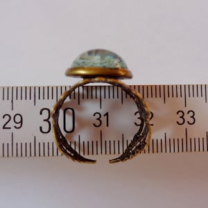 May include: A gold-toned adjustable ring with a blue and white glass cabochon. The ring is shown on a ruler, measuring approximately 30 mm in diameter.