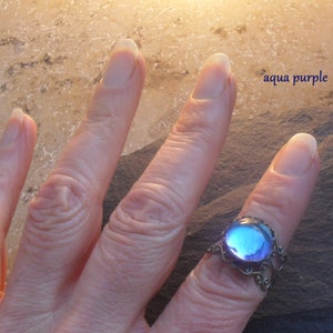 Op de afbeelding: Een ring met een grote, ronde, iriserende blauwe en paarse glazen steen, gezet in een zilveren filigraanontwerp. De ring zit aan een vinger.
