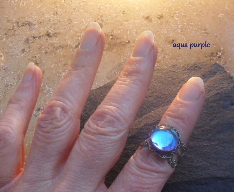 Op de afbeelding: Een ring met een grote, ronde, iriserende blauwe en paarse glazen steen, gezet in een zilveren filigraanontwerp. De ring zit aan een vinger.