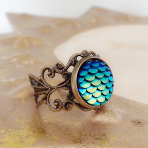 Op de afbeelding: Een ring met een blauw en groen iriserend zeemeermin-schubbenontwerp, gezet in een sierlijke bronzen filigraansetting.