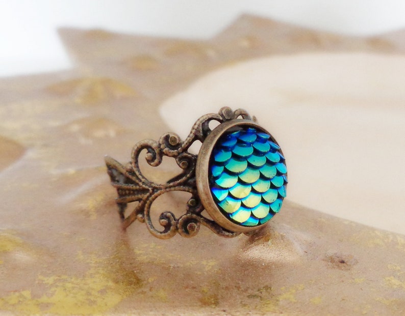 Op de afbeelding: Een ring met een blauw en groen iriserend zeemeermin-schubbenontwerp, gezet in een sierlijke bronzen filigraansetting.