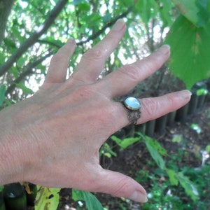 May include: A silver ring with a white oval gemstone set in a filigree design. The ring is worn on a person's finger.