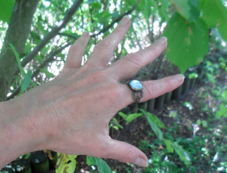 May include: A silver ring with a white oval gemstone set in a filigree design. The ring is worn on a person's finger.