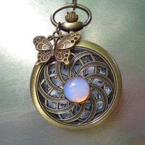 May include: Antique bronze pocket watch with a butterfly charm. The watch face features a swirling, openwork design with a light blue, iridescent stone in the center. The watch has a chain and a loop for hanging.