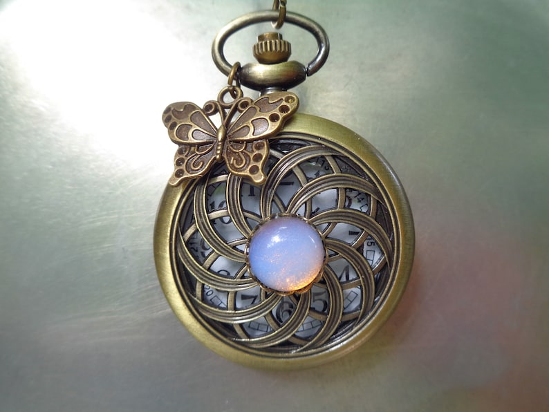 May include: Antique bronze pocket watch with a butterfly charm. The watch face features a swirling, openwork design with a light blue, iridescent stone in the center. The watch has a chain and a loop for hanging.